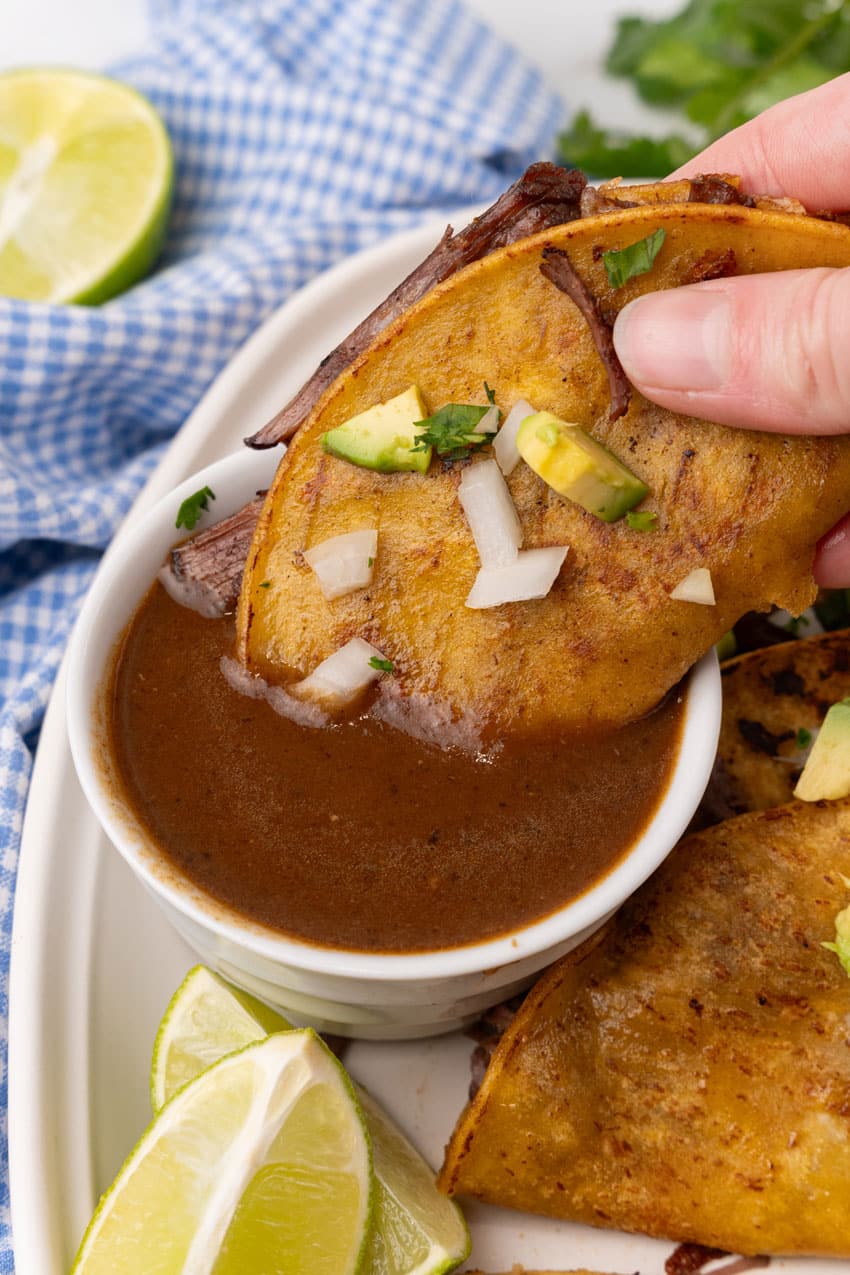 a hand dipping a quesa birria taco in a small white bowl of birria sauce