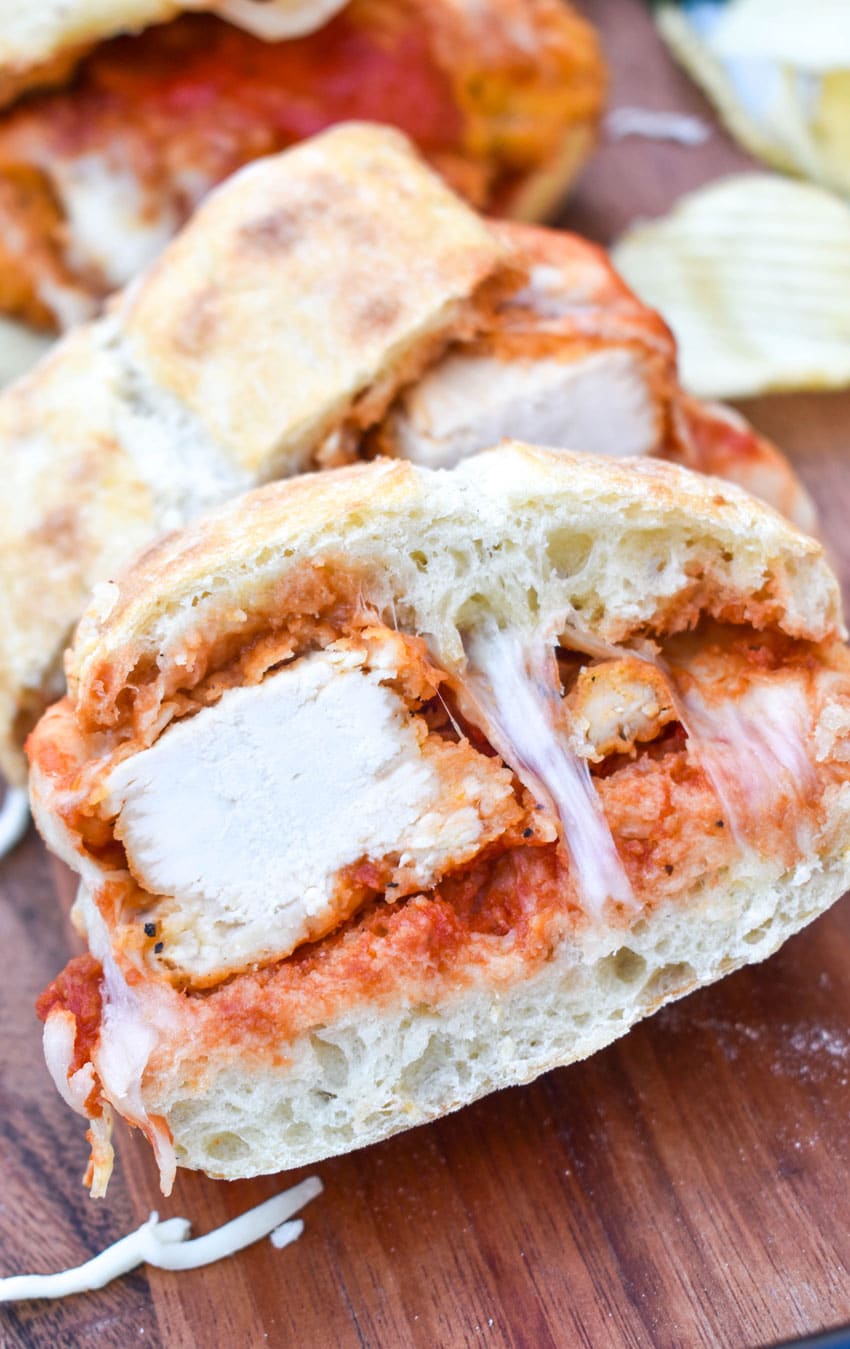 a chicken parmesan sandwich on a wooden cutting board with fresh herbs and crispy potato chips in the background