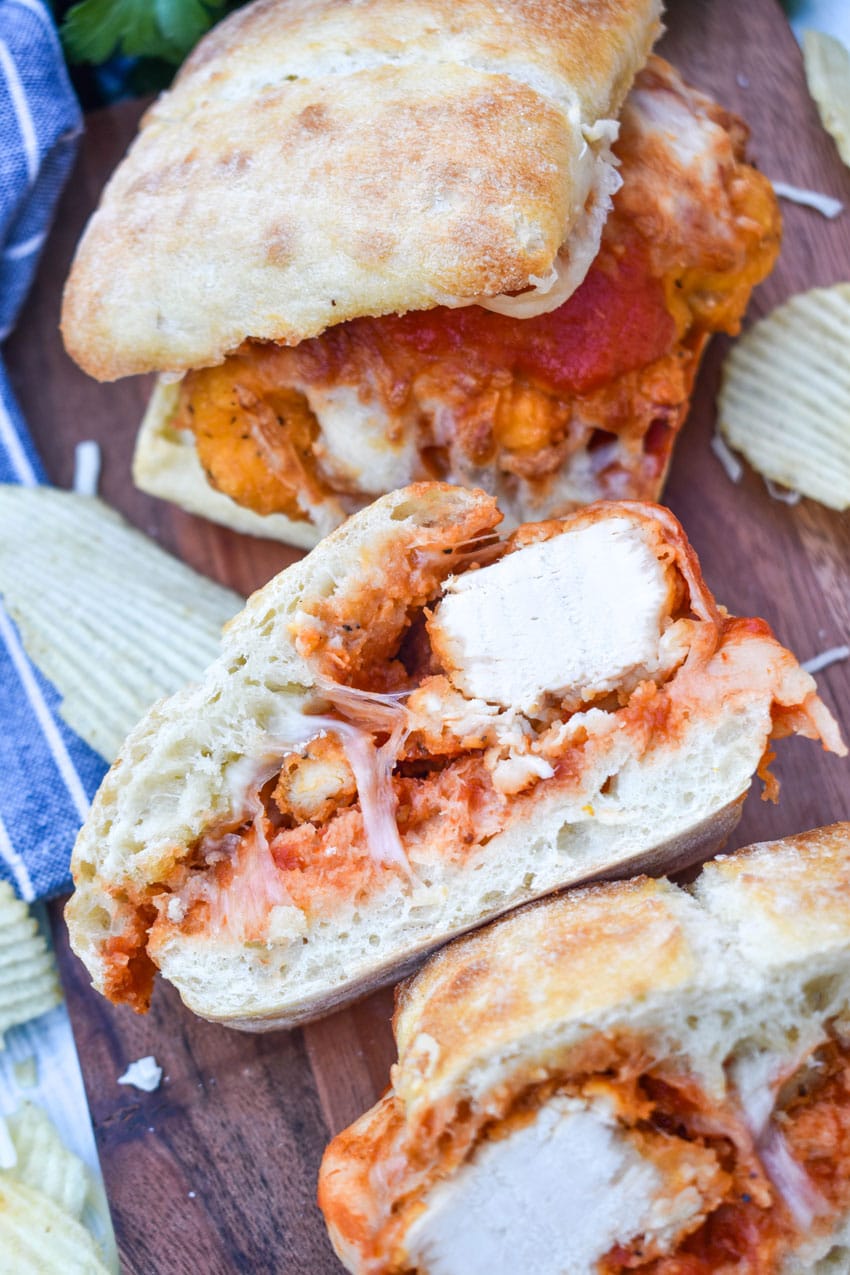 a chicken parmesan sandwich on a wooden cutting board with fresh herbs and crispy potato chips in the background
