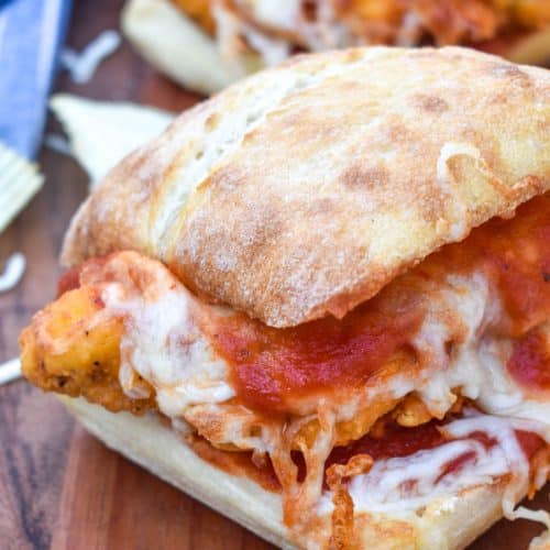 a chicken parmesan sandwich on a wooden cutting board with fresh herbs and crispy potato chips in the background