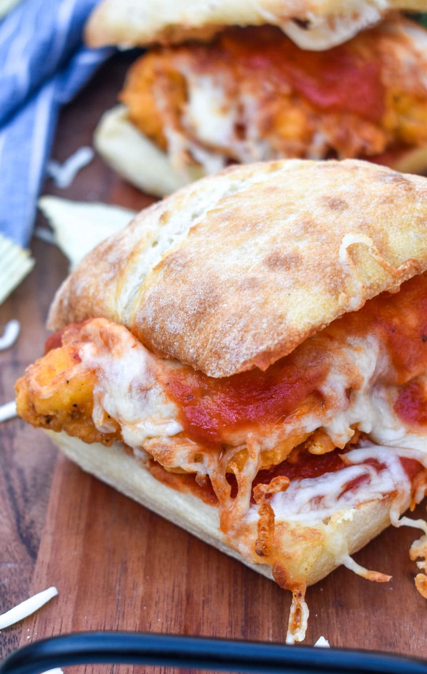 a chicken parmesan sandwich on a wooden cutting board with fresh herbs and crispy potato chips in the background