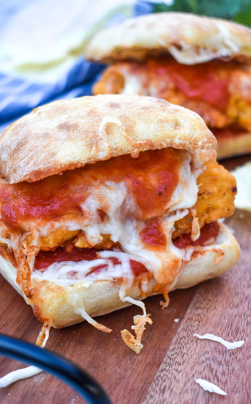 a chicken parmesan sandwich on a wooden cutting board with fresh herbs and crispy potato chips in the background
