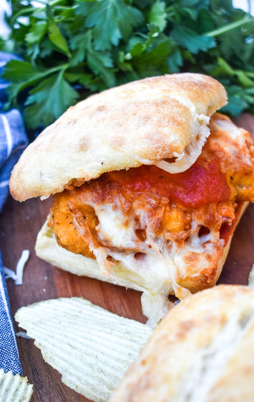 a chicken parmesan sandwich on a wooden cutting board with fresh herbs and crispy potato chips in the background