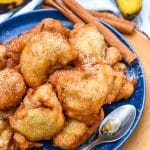 a pile of fried banana fritters topped with powdered sugar and syrup on a blue plate with a small spoon on the side