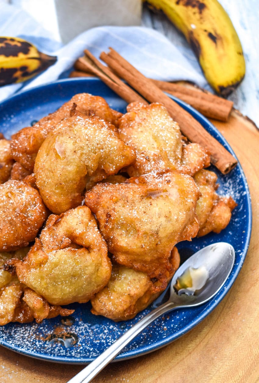 a pile of fried banana fritters topped with powdered sugar and syrup on a blue plate with a small spoon on the side