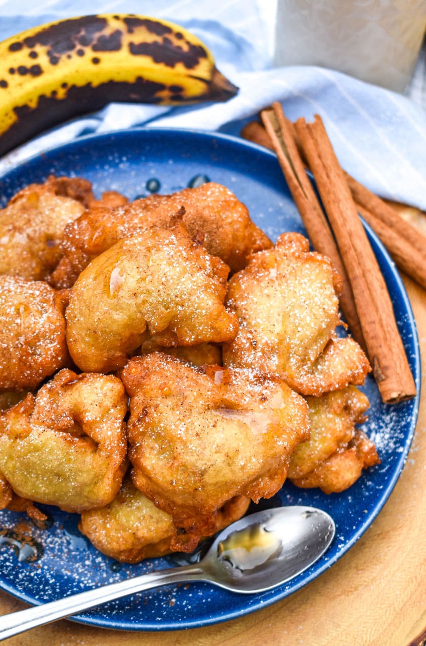 a pile of fried banana fritters topped with powdered sugar and syrup on a blue plate with a small spoon on the side