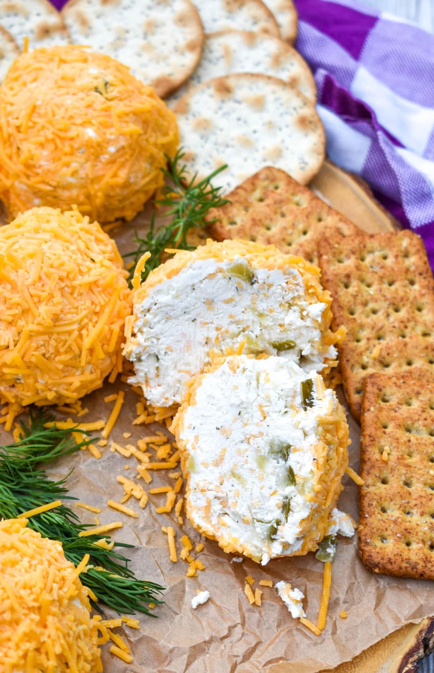 TWO HALVES OF A MINI DILL PICKLE CHEESE BALL ON A WOODEN CUTTING BOARD SURROUNDED BY ASSORTED CRACKERS
