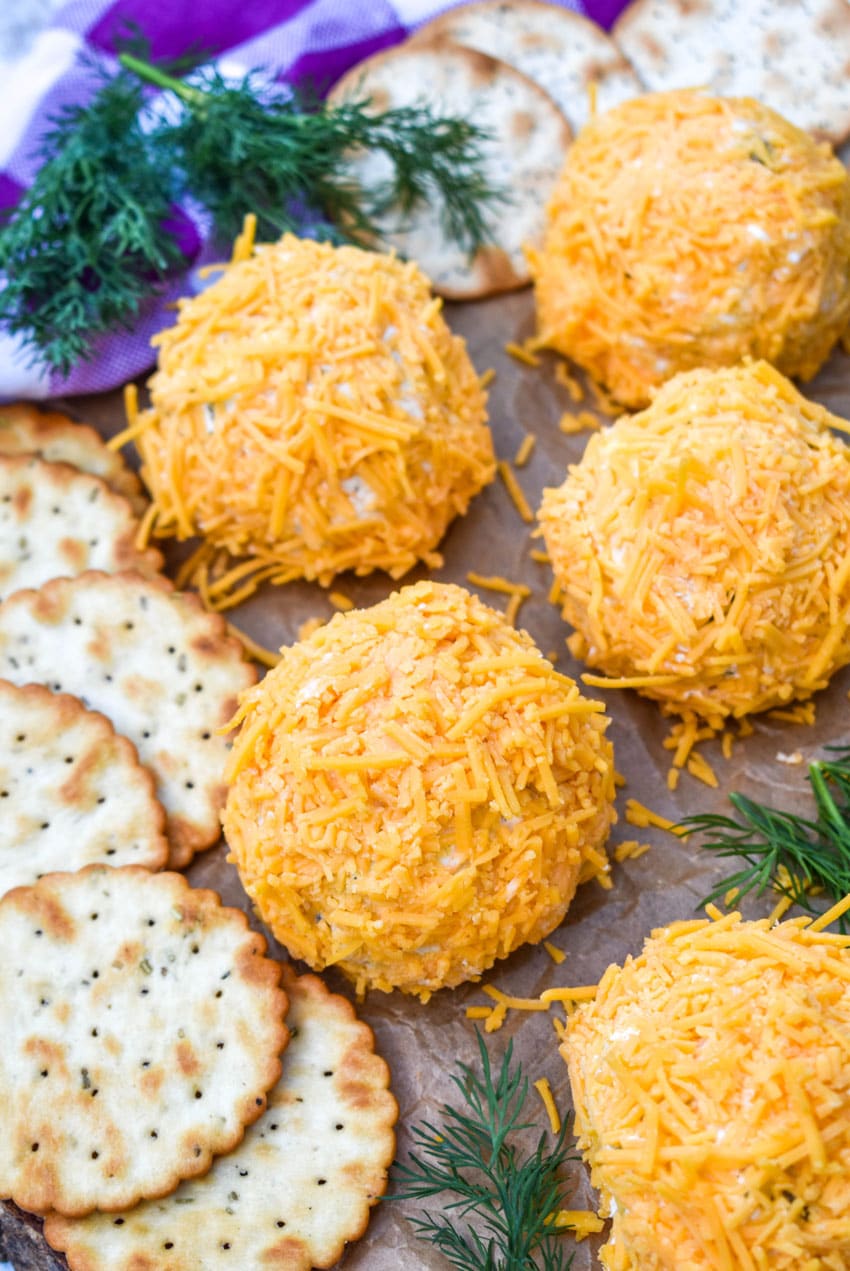 MINI DILL PICKLE CHEESE BALLS ON A WOODEN CUTTING BOARD WITH CRACKERS AND SPRIGS OF FRESH DILL ON THE SIDE