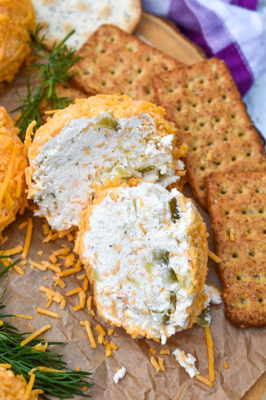 TWO HALVES OF A MINI DILL PICKLE CHEESE BALL ON A WOODEN CUTTING BOARD SURROUNDED BY ASSORTED CRACKERS