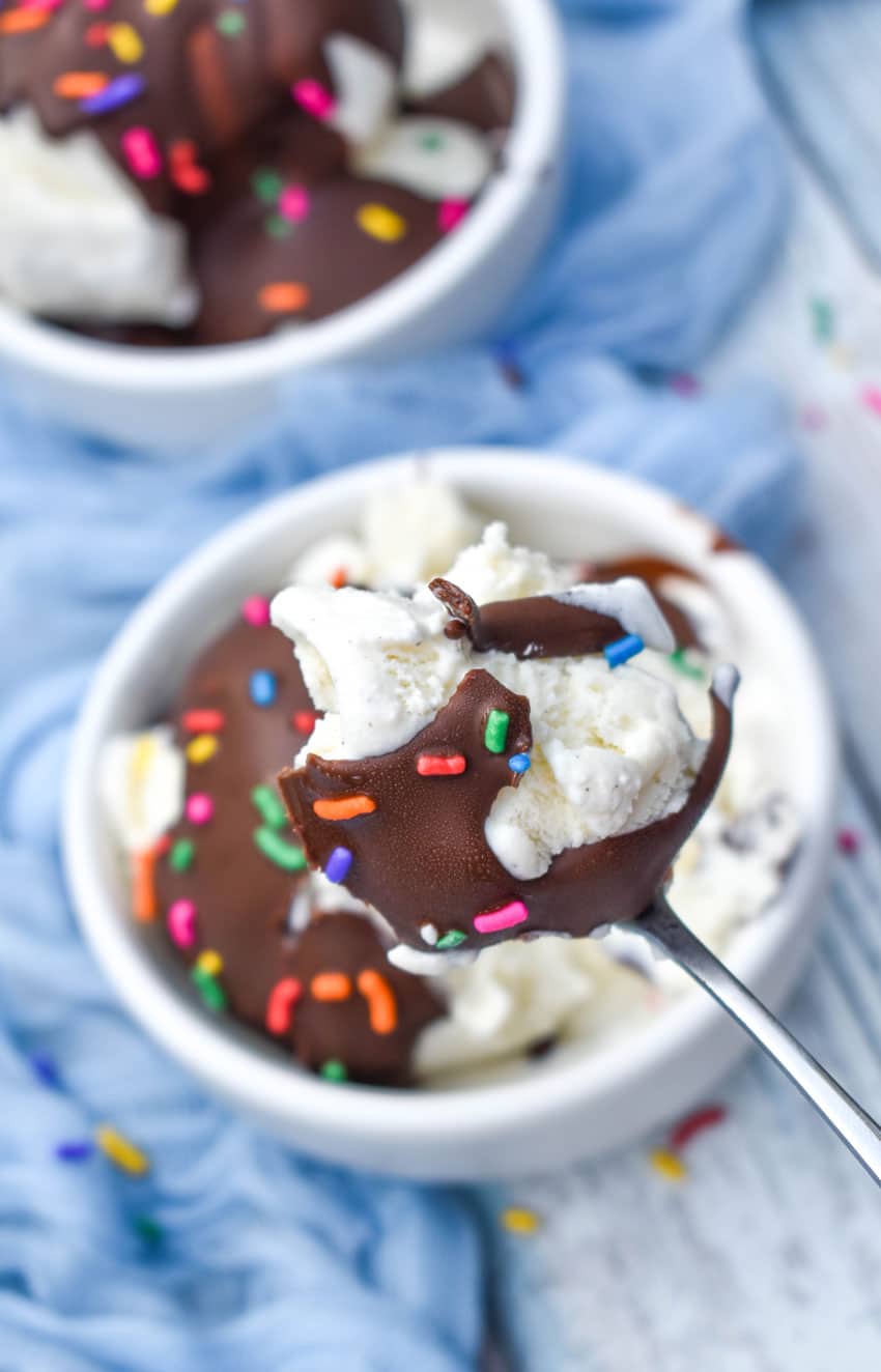 a silver spoon holding up a scoop of vanilla ice cream coated in hardened chocolate with rainbow sprinkles on top