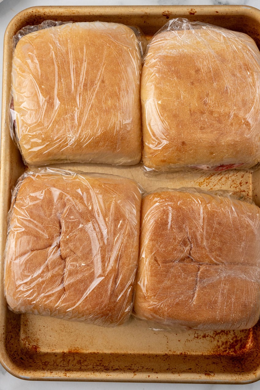 plastic wrapped pressed italian picnic sandwiches on a baking sheet