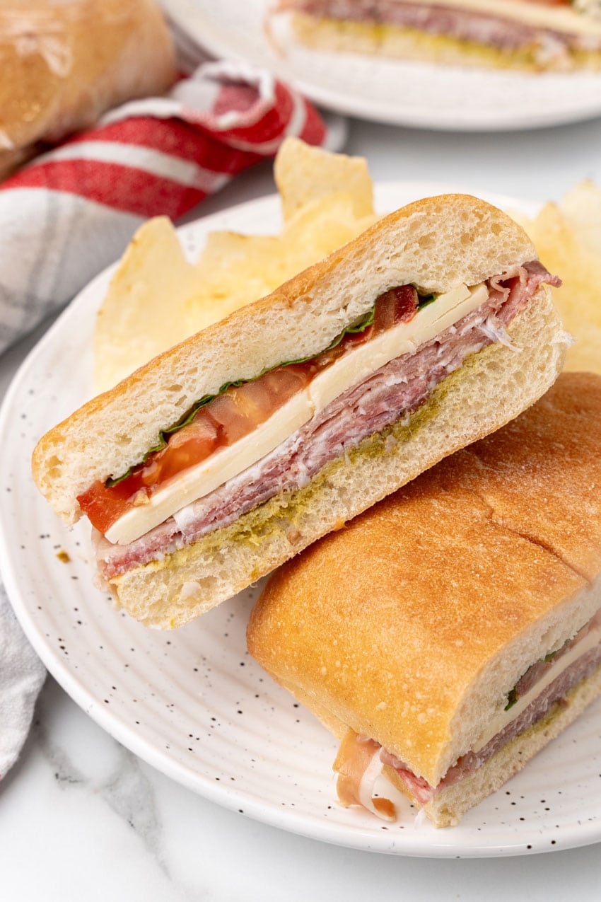 two halves of an italian pressed picnic sandwich stacked together on a white plate with potato chips on the side