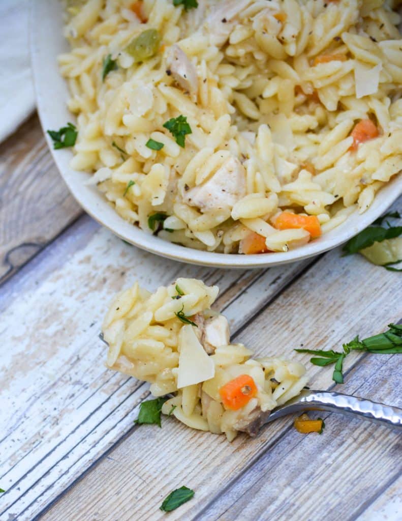 a silver fork holding a scoop of creamy chicken and orzo pasta resting on a wooden table