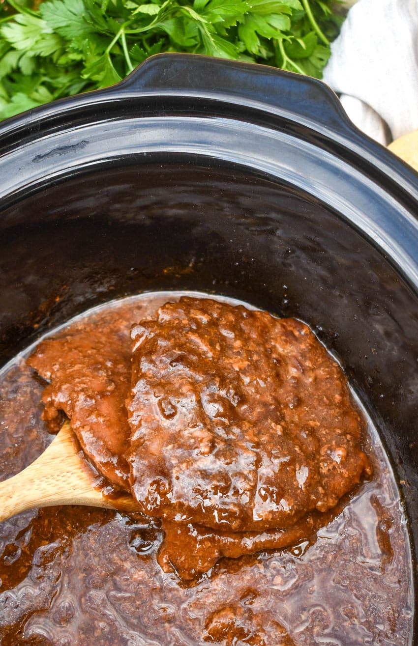 a wooden spatula scooping slow cooker cube steaks covered in gravy out of a black crockpot