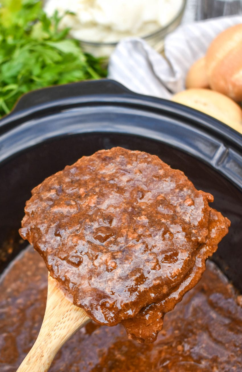a wooden spatula holding up a piece of slow cooker cube steak with gravy above a crockpot