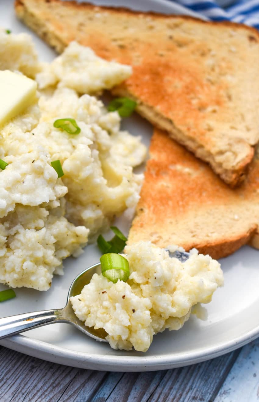 a silver spoon holding a scoop of creamy crockpot grits resting on the edge of a white plate with slices of toast on the side