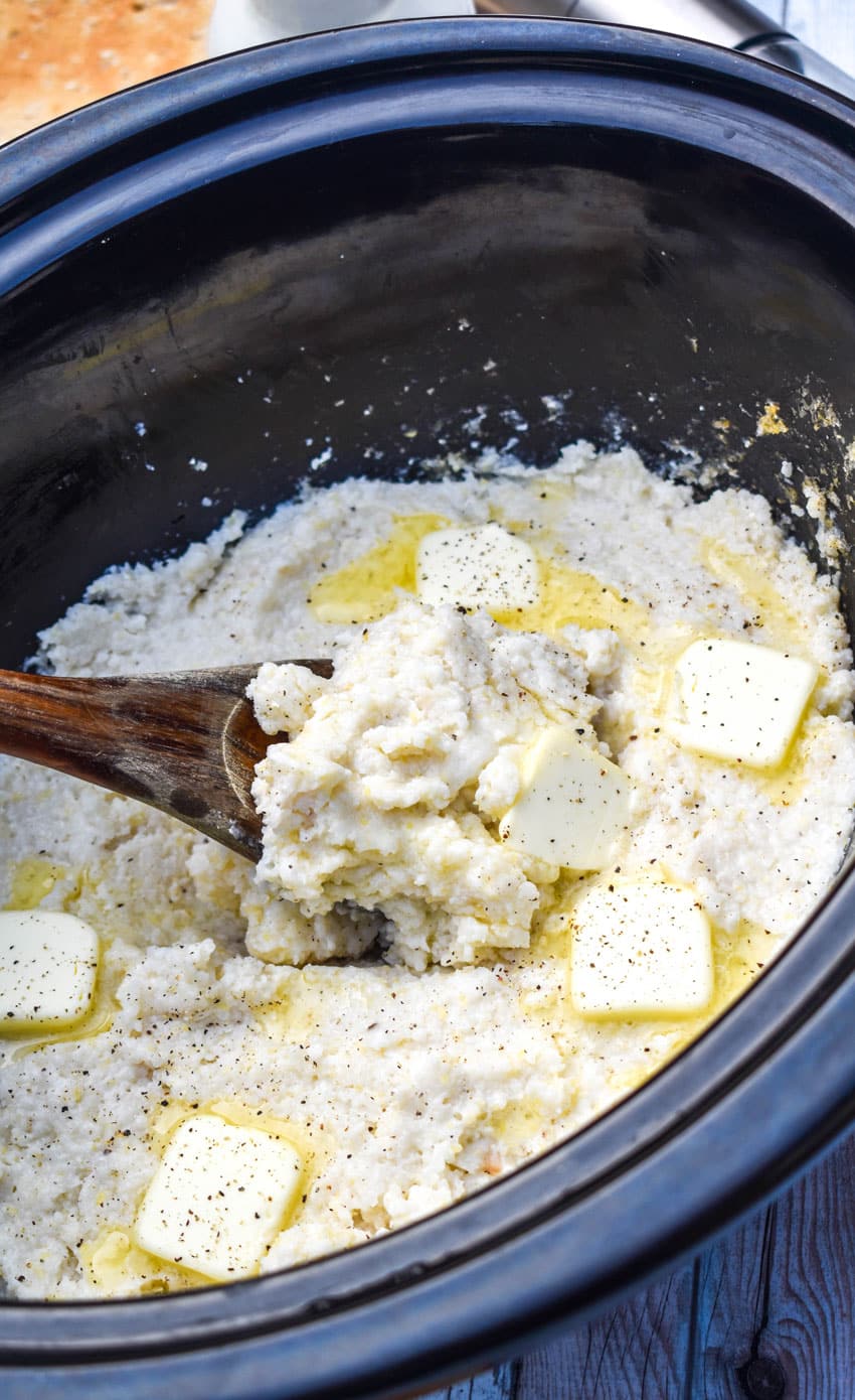 a wooden spoon scooping creamy grits topped with butter out of the bowl of a slow cooker