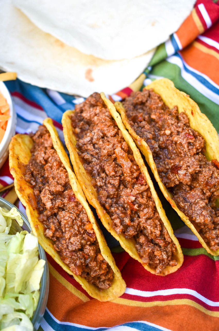 slow cooker taco meat in three crispy tortilla shells arranged in a row on a cloth napkin
