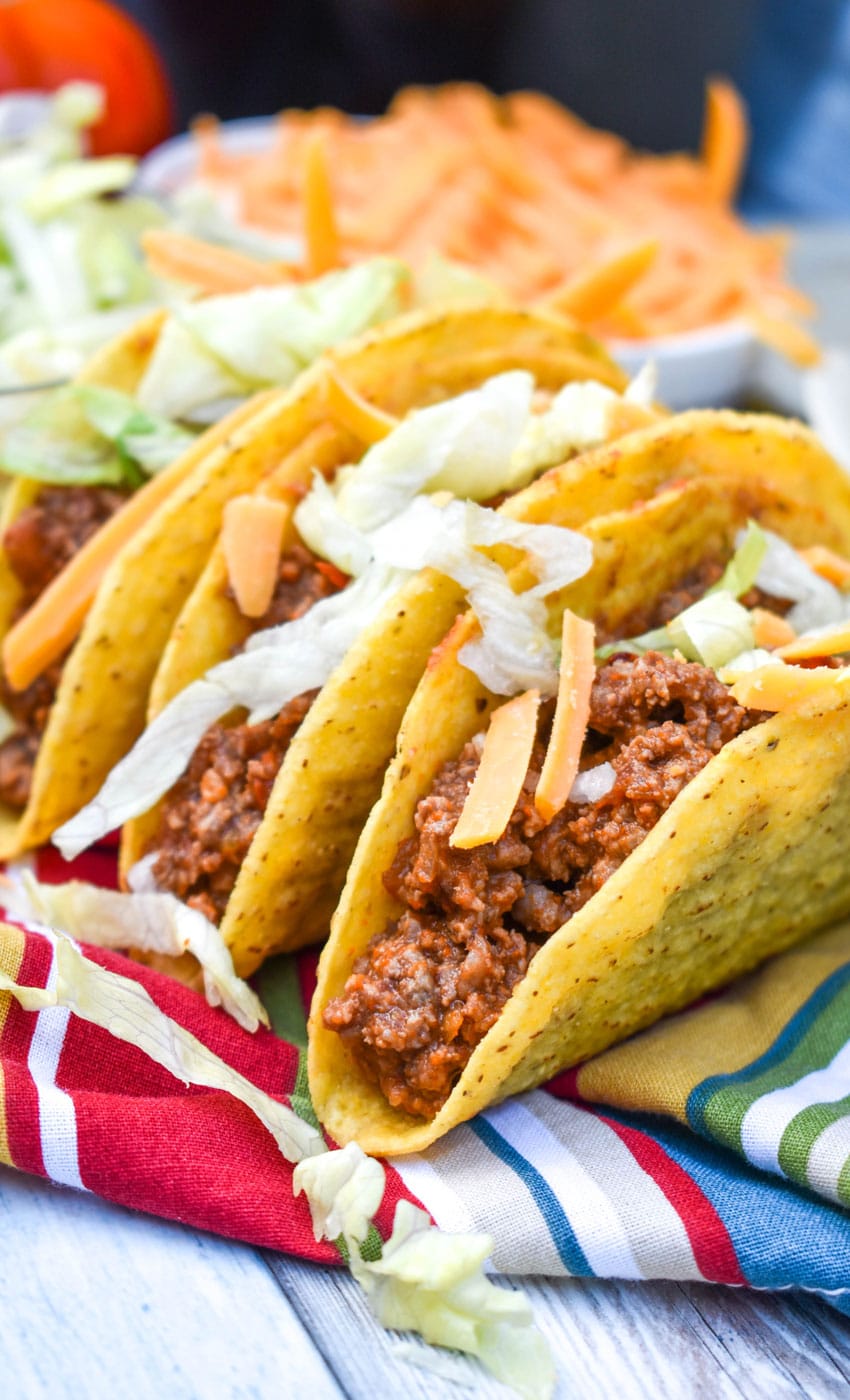 slow cooker taco meat in three crispy tortilla shells arranged in a row on a cloth napkin