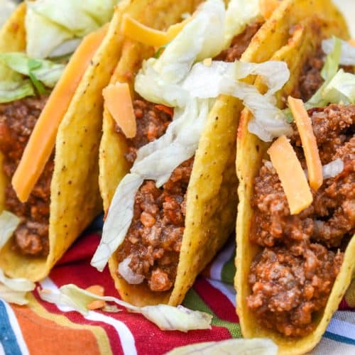 slow cooker taco meat in three crispy tortilla shells arranged in a row on a cloth napkin