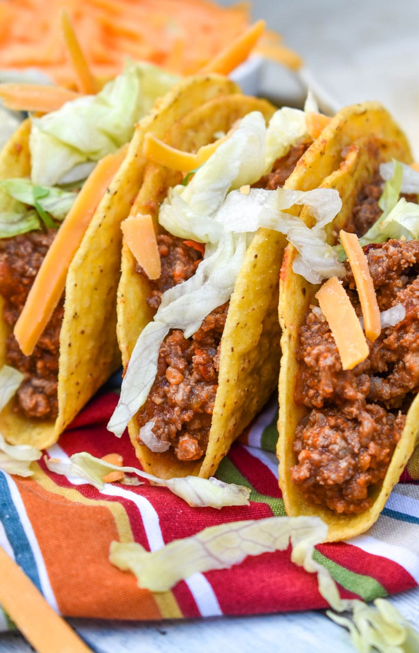 slow cooker taco meat in three crispy tortilla shells arranged in a row on a cloth napkin