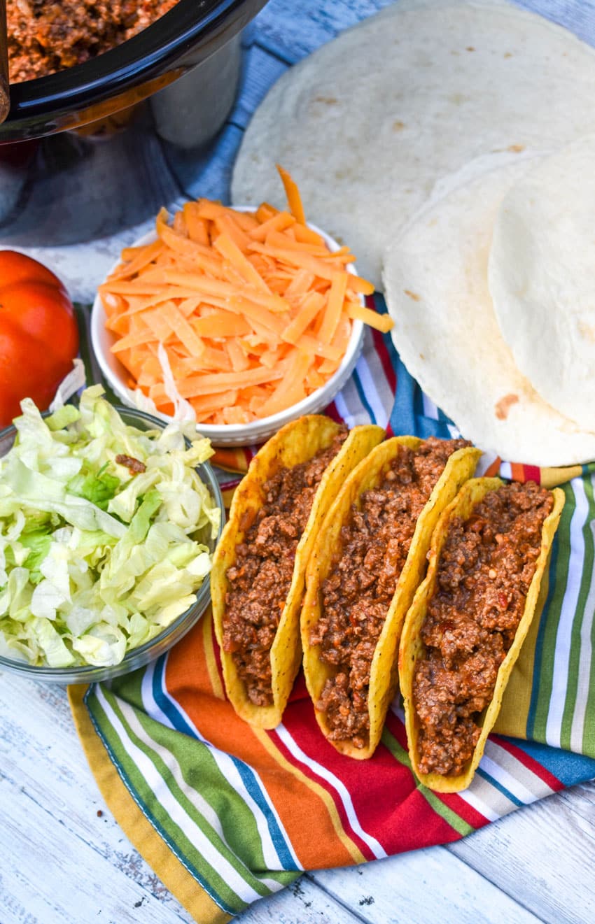 slow cooker taco meat in three crispy tortilla shells arranged in a row on a cloth napkin