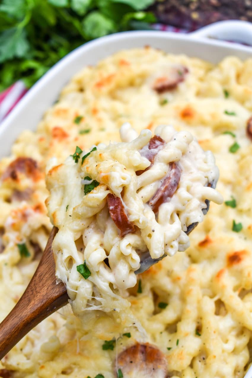 a wooden spoon holding up a scoop of baked alfredo pasta with sausage