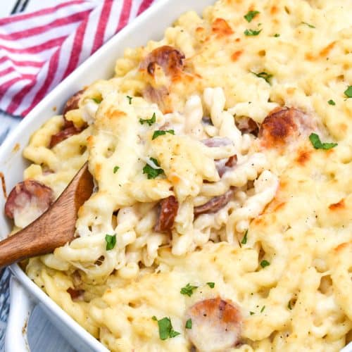 a wooden spoon scooping baked alfredo pasta with sausage out of a white casserole dish