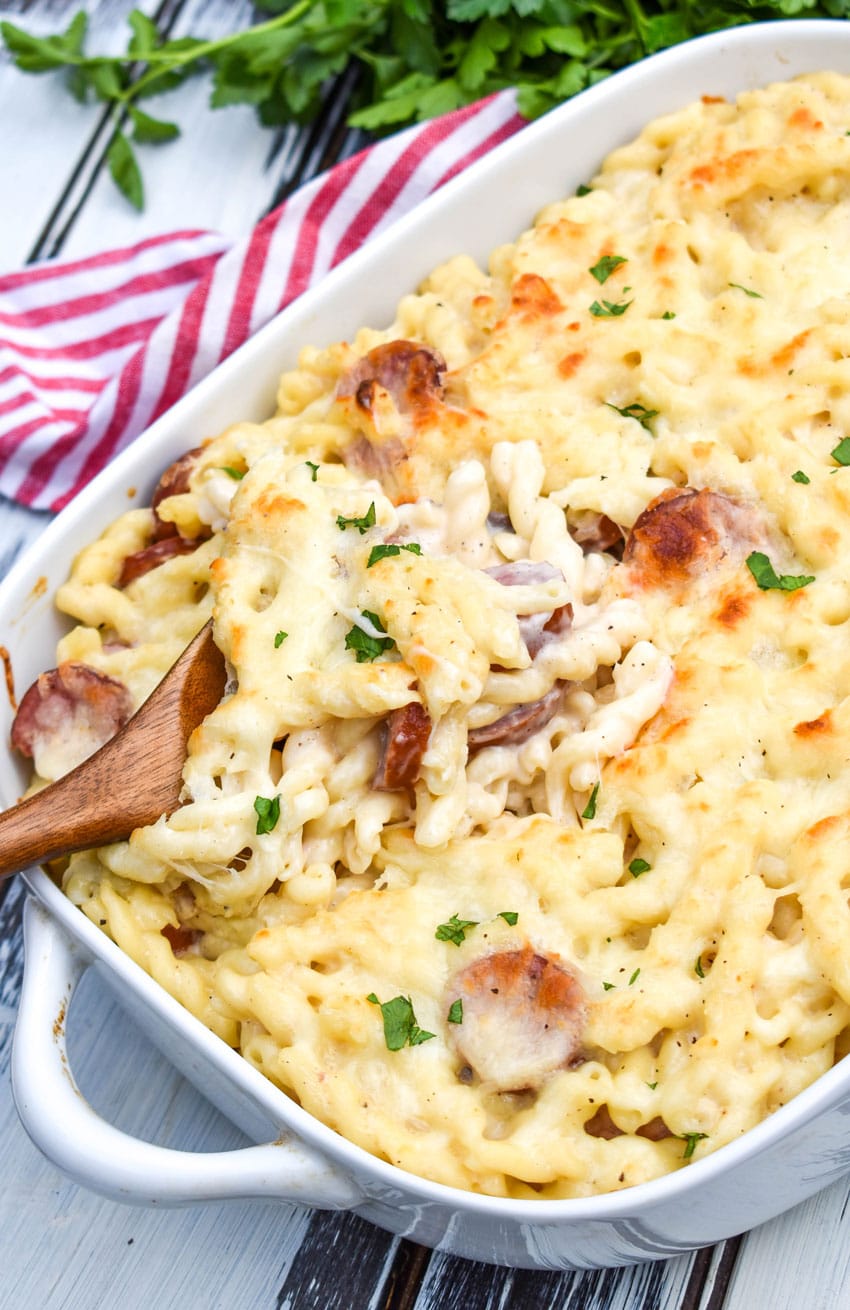 Baked Alfredo Pasta With Sausage a wooden spoon scooping baked alfredo pasta with sausage out of a white casserole dish