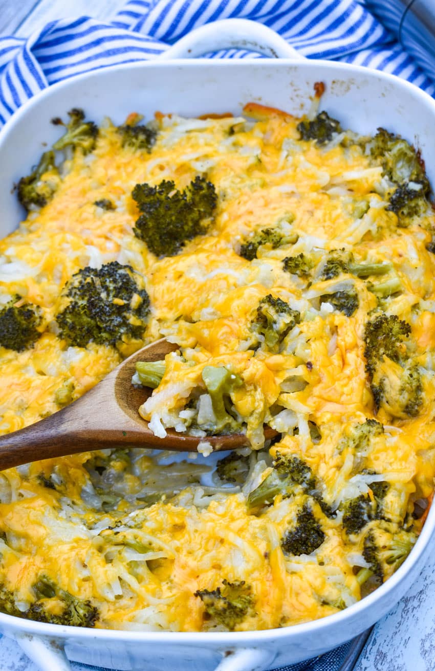 a wooden spoon scooping cheesy broccoli potato casserole out of a white baking dish