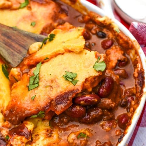 a wooden spoon scooping chili cornbread casserole out of a white baking dish