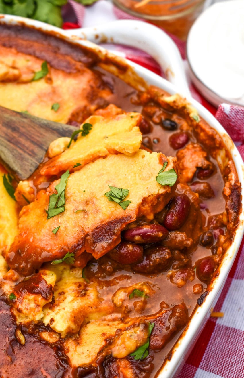 a wooden spoon scooping chili cornbread casserole out of a white baking dish