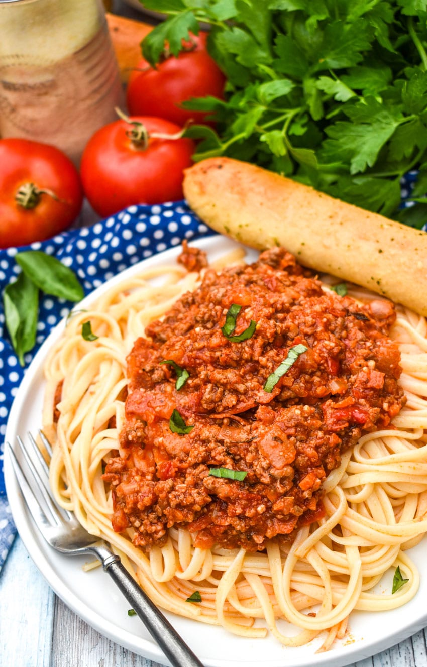 OLIVE GARDEN MEAT SAUCE RECIPE OVER COOKED SPAGHETTI ON A WHITE PLATE WITH A SILVER FORK AND A BREAD STICK ON THE SIDES