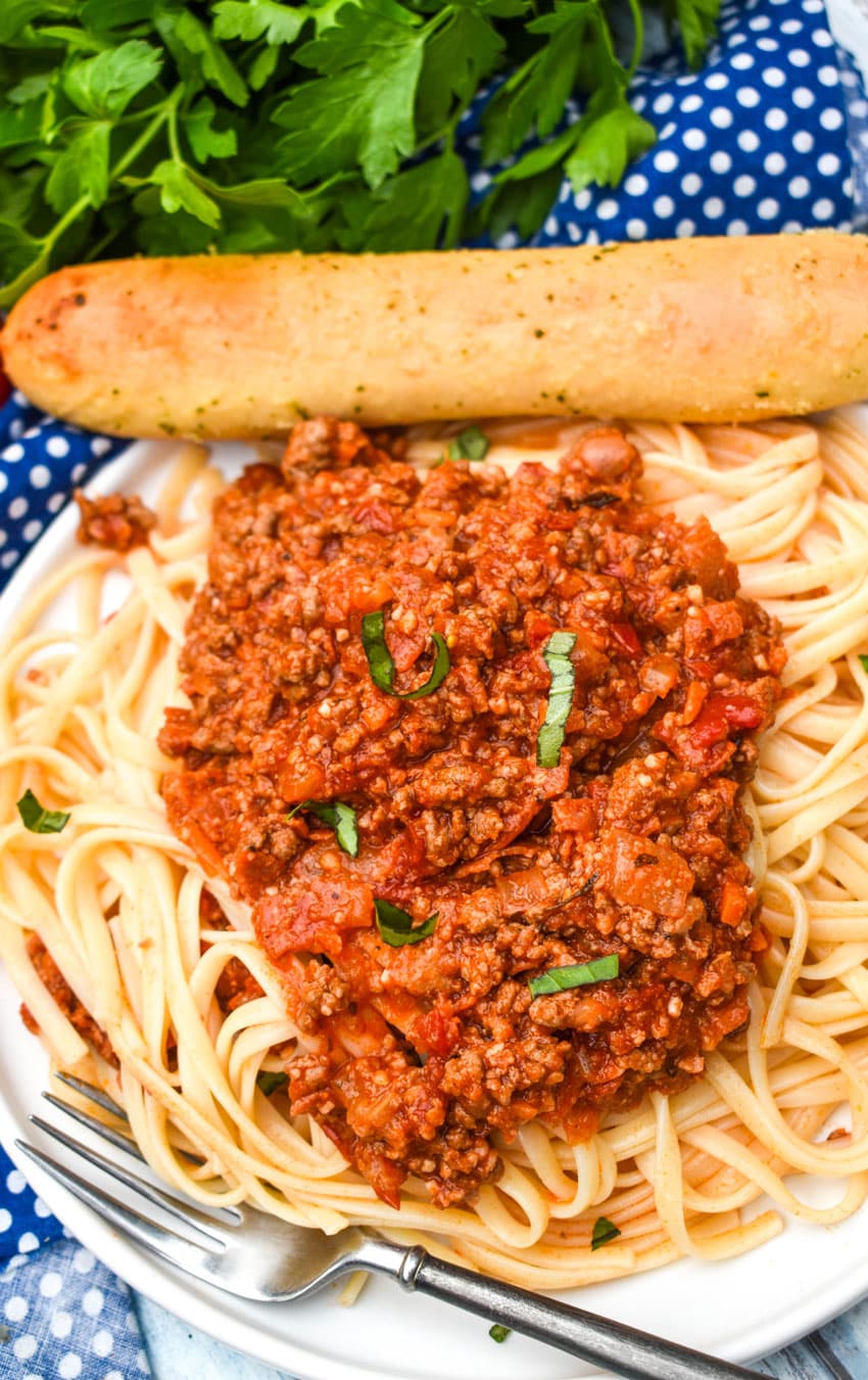 HOMEMADE MEAT SAUCE OVER COOKED SPAGHETTI ON A WHITE PLATE WITH A SILVER FORK AND A BREAD STICK ON THE SIDES