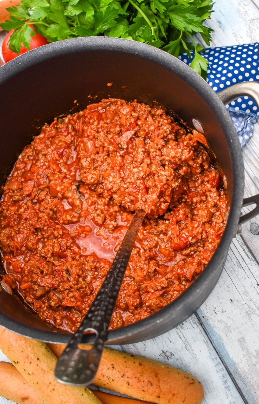 A SILVER LADLE SCOOPING THREE MEAT PASTA SAUCE OUT OF A BLACK POT