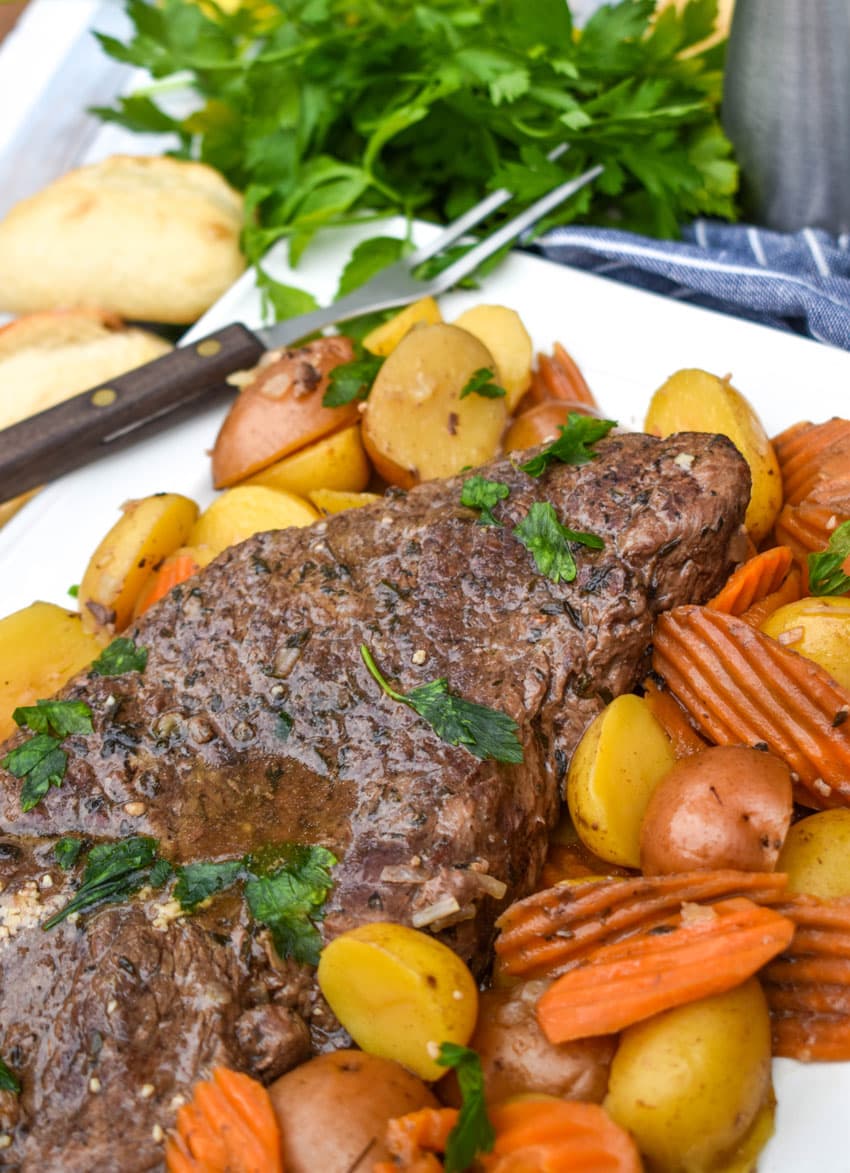 THE BEST SLOW COOKER POT ROAST RECIPE SURROUNDED BY COOKED CARROTS AND POTATOES ON A WHITE PLATTER