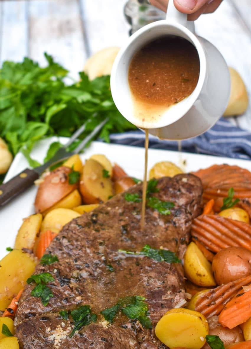 BROWN SAUCE BEING POURED OVER A CROCKPOT POT ROAST ON A WHITE PLATTER SURROUNDED BY COOKED POTATOES AND CARROTS