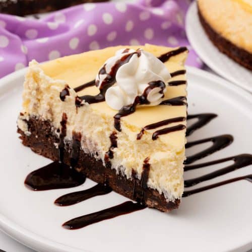 a slice of brownie bottom cheesecake topped with chocolate sauce and a spritz of whipped cream on a white plate