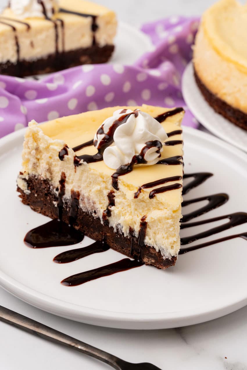 Brownie Bottom Cheesecake a slice of brownie bottom cheesecake topped with chocolate sauce and a spritz of whipped cream on a white plate