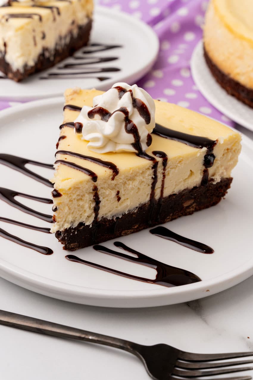 a slice of brownie bottom cheesecake topped with chocolate sauce and a spritz of whipped cream on a white plate