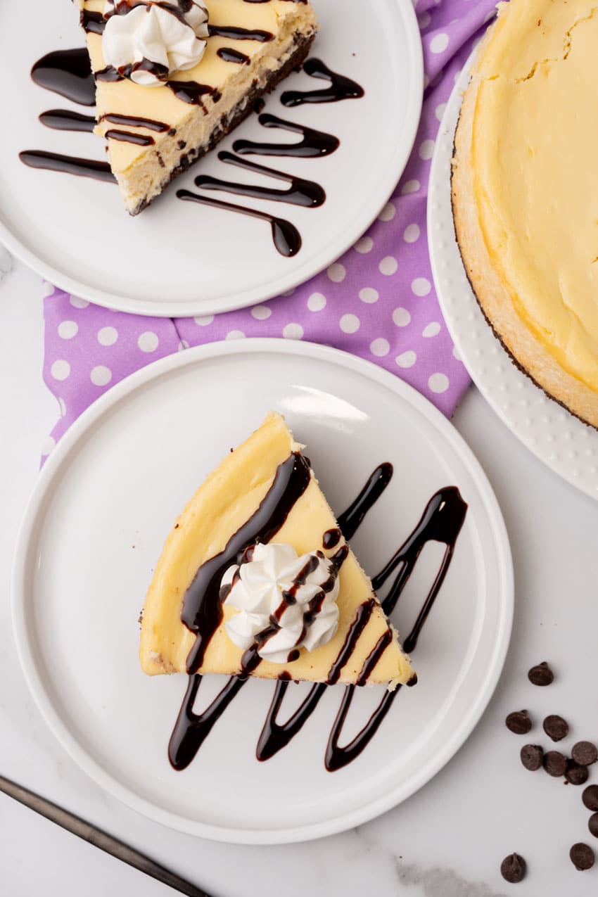 slices of brownie bottom cheesecake topped with chocolate sauce and a spritz of whipped cream on two white plates