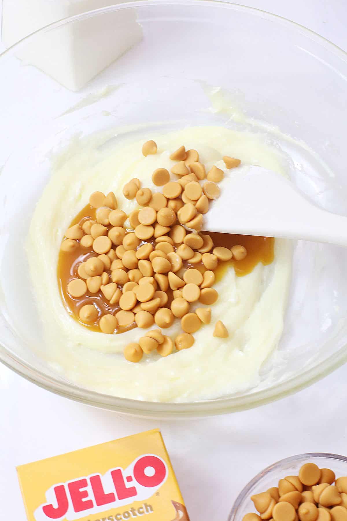 butterscotch chips and butterscotch syrup being folded into cheesecake filling in a glass mixing bowl