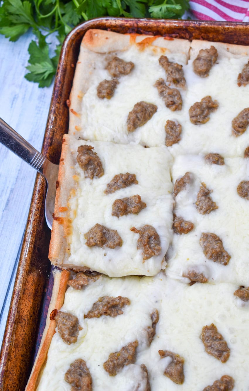 a silver spatula scooping a slice of cafeteria breakfast pizza off a metal baking sheet
