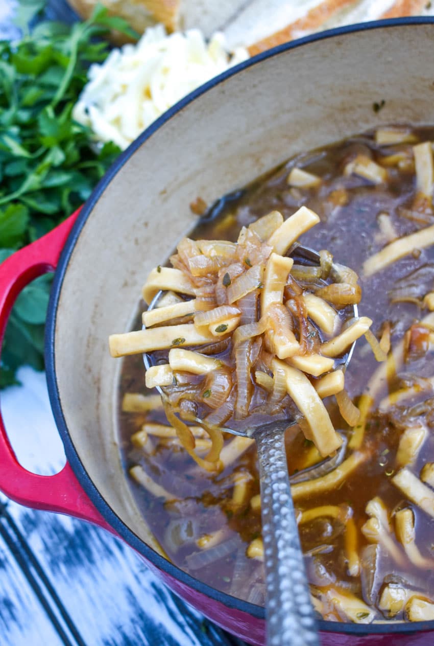 a silver ladle scooping french onion noodle soup out of a large dutch oven