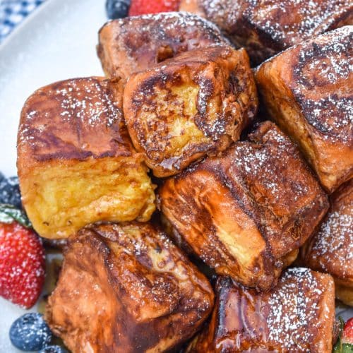 hawaiian roll french toast sprinkled with powdered sugar stacked on a white plate and drizzled with syrup with fresh berries on the side