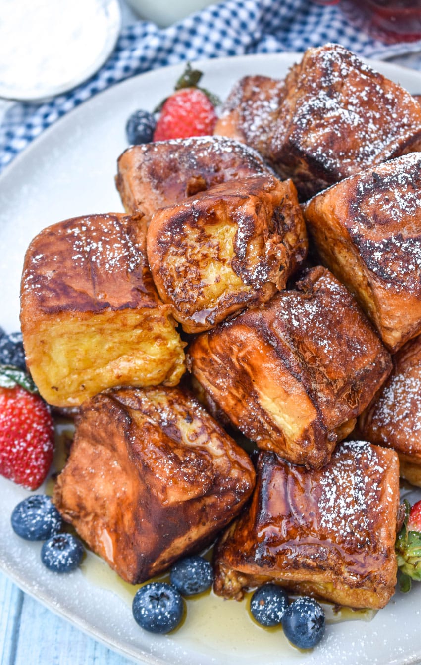 hawaiian roll french toast sprinkled with powdered sugar stacked on a white plate and drizzled with syrup with fresh berries on the side