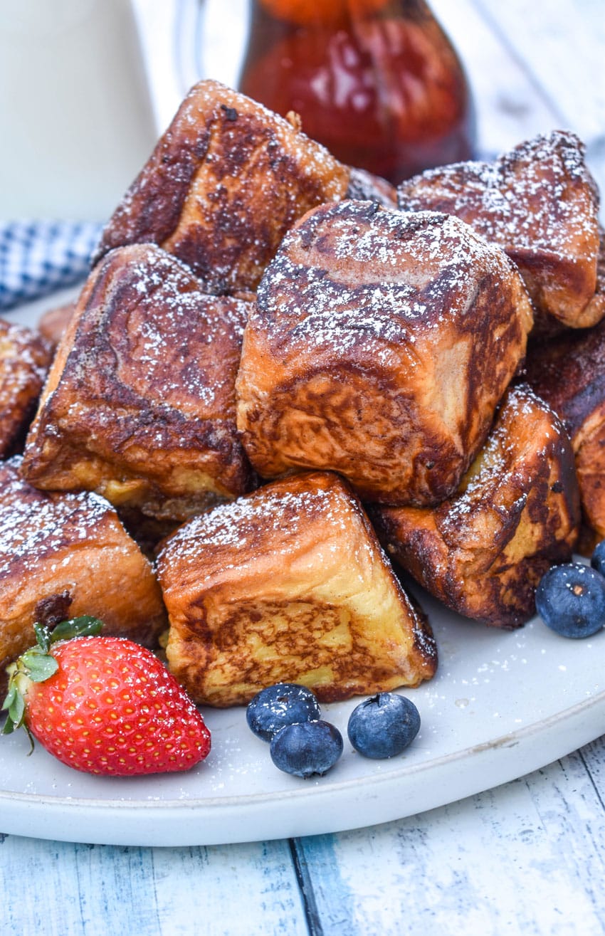 hawaiian roll french toast sprinkled with powdered sugar stacked on a white plate with fresh berries on the side