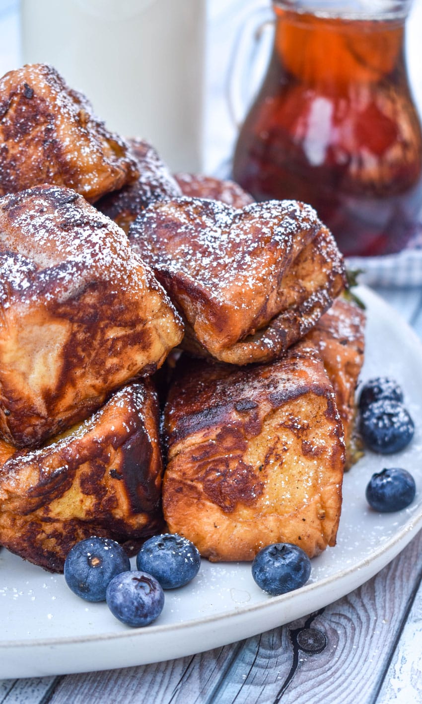 hawaiian roll french toast sprinkled with powdered sugar stacked on a white plate with fresh berries on the side