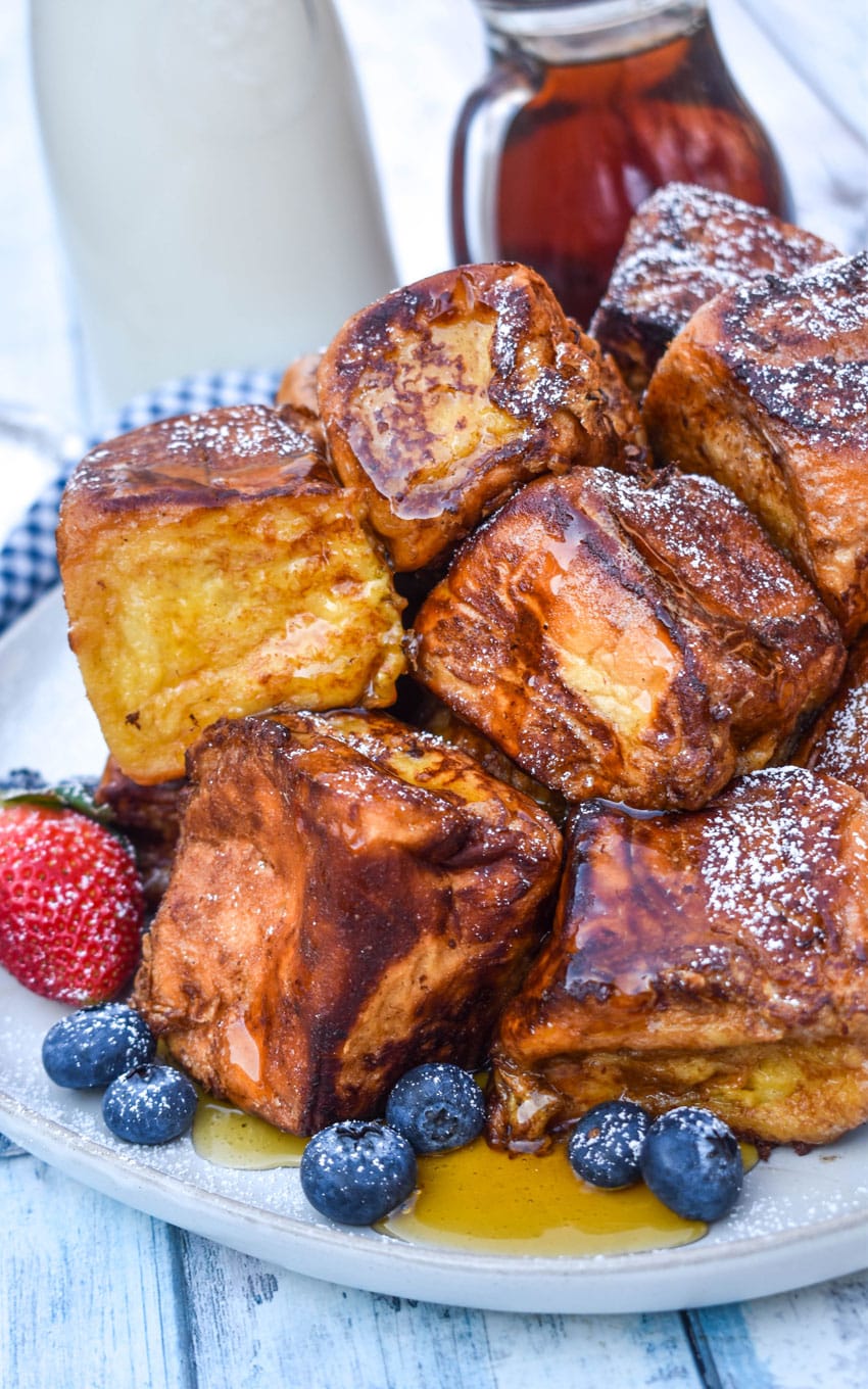 hawaiian roll french toast sprinkled with powdered sugar stacked on a white plate and drizzled with syrup with fresh berries on the side