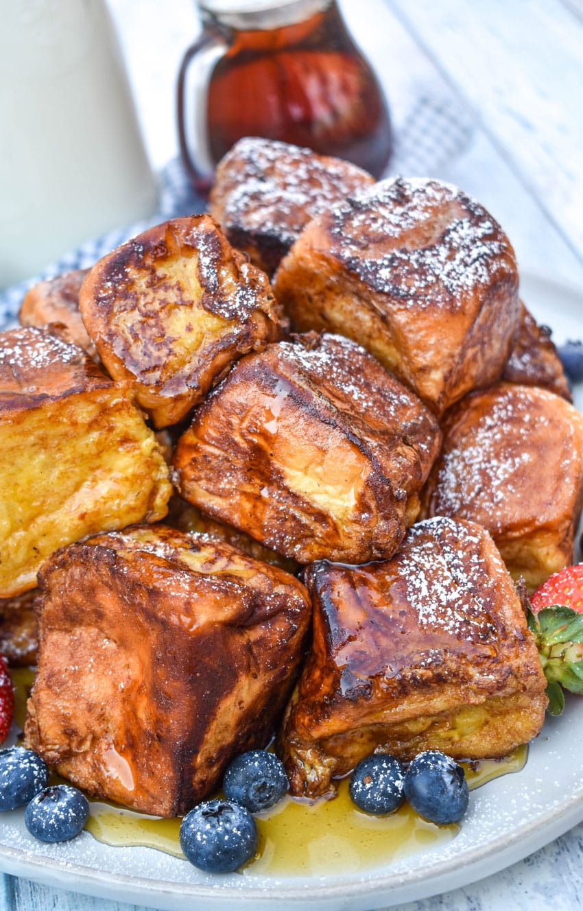 hawaiian roll french toast sprinkled with powdered sugar stacked on a white plate and drizzled with syrup with fresh berries on the side